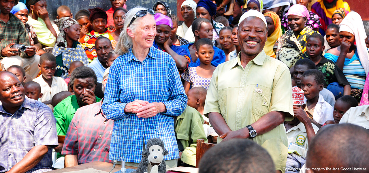 Jane Goodall in a blue shirt standing outside beside a smiling man in a green shirt in the middle of a large colorful crowd of people 