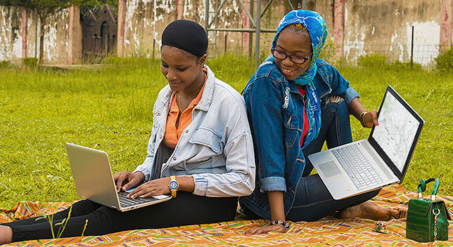 Two casually dressed people sitting back-to-back in a grassy field, each with a laptop in their laps, the one on the right peering over her shoulder to speak to the other