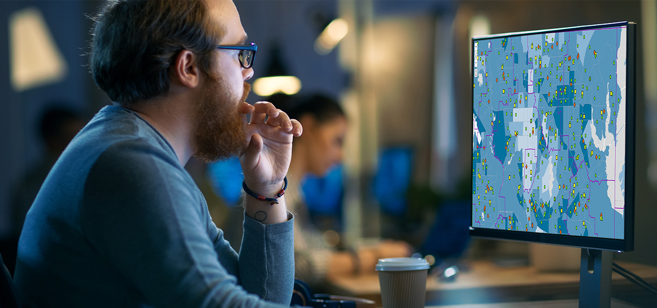 Una persona con una maglietta blu a maniche lunghe siede a una scrivania in uno spazio ufficio condiviso, utilizzando un monitor per accedere a una mappa digitale