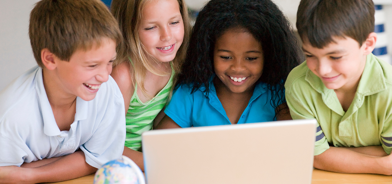 Four smiling children grouped closely around a shared laptop in a brightly lit classroom