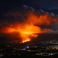 An aerial view of a city brightly lit for nighttime with a raging fire in the middle sending vivid orange and yellow-lit smoke into a dark night sky