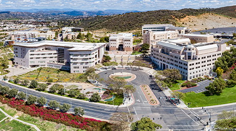 Aerial photo of a modern university with white buildings against a backdrop of rolling brown hills under a cloudy blue sky