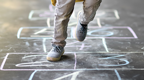 A closeup photo of a child’s legs and feet in mid-leap on a chalk hopscotch grid