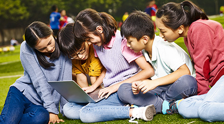 A family with several children and adults groups closely together around a shared laptop while sitting in the grass in a sunny park