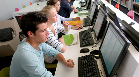 Four students seated in a row looking at spreadsheets of data at matching computer stations in a school computer lab