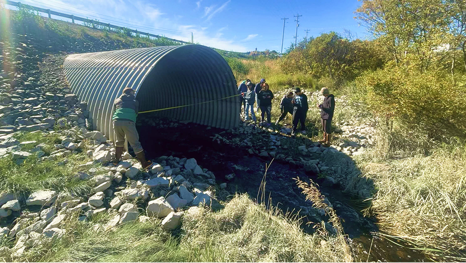 A group of students standing outside a large drainage pipe in a sunny field