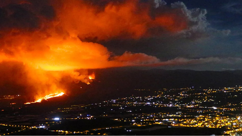An aerial view of a city brightly lit for nighttime with a raging fire in the middle sending vivid orange and yellow-lit smoke into a dark night sky