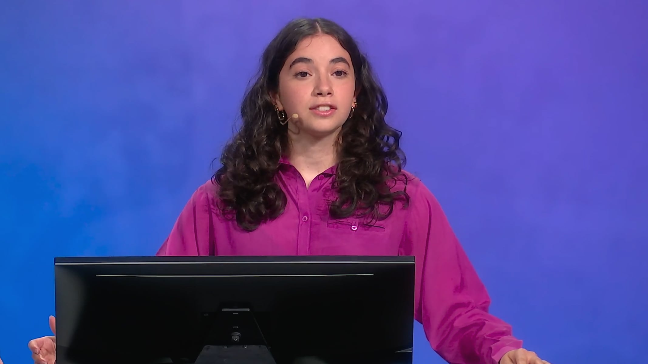 A student wearing a bright pink collared shirt speaking onstage 