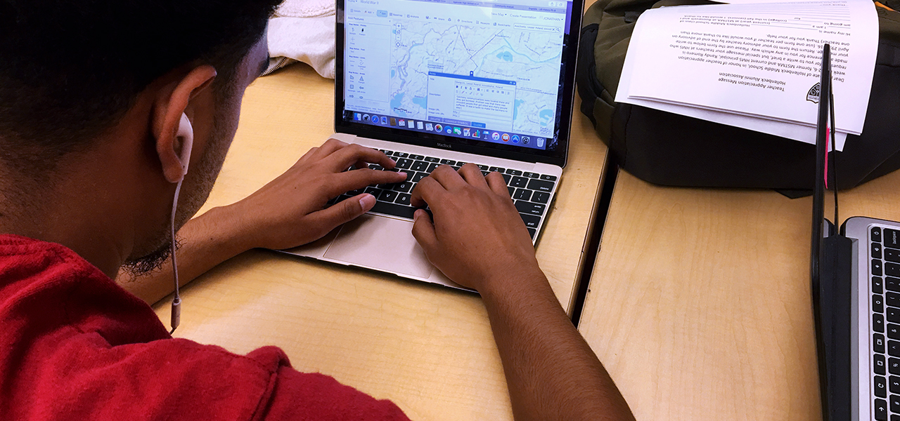 A student wearing earbuds and a red tee shirt sitting at a classroom desk accessing a digital map on a small laptop