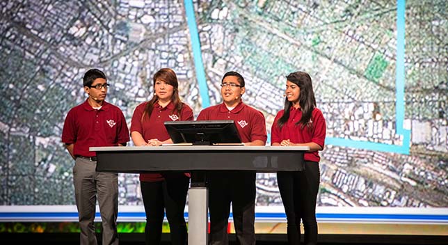 Four people in red polo shirts standing at a large podium onstage in front of a large aerial photo of a dense city