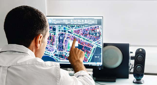 A person in a white collared shirt using a desktop computer to view a digital city map in blue, white, and purple
