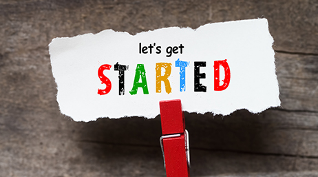 A red clothespin holding a ragged scrap of white paper printed with the multicolored words “let’s get started” against a rough wood background