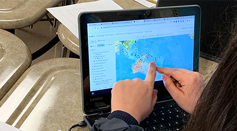 A student sitting at a classroom desk points to a digital map on a small laptop