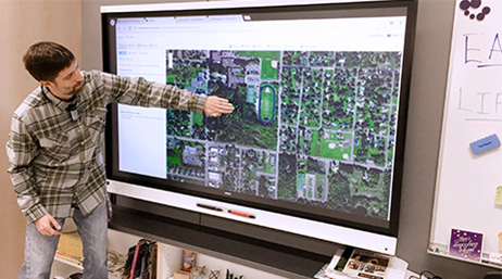 A student gestures to a large wall-mounted screen displaying a digital map at the front of a classroom
