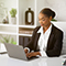 A smiling person dressed in a neat white collared shirt and dark blazer using a small laptop in a sunlit modern white office