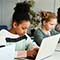 A row of four young students studying on laptops and tablets in a modern classroom with white walls and tables