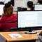 A photo taken from the back of a classroom full of students seated at rows of wood-toned desks, each with a computer station in front of them