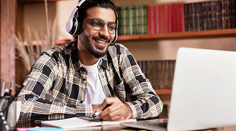 A photo of a smiling student in a blue and white plaid shirt and a headset, seated at a desk looking at an open laptop in a library setting