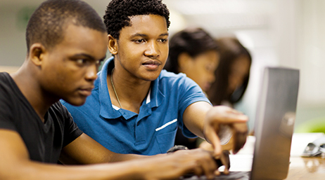 Foto von zwei leger gekleideten Studierenden einer Hochschule an einem Tisch in einem sanft beleuchteten Klassenzimmer, die gemeinsam einen geöffneten Laptop nutzen und die Monitoranzeige besprechen