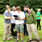 A photo of a small crowd of casually-dressed college students standing in a green tree-lined field, some studying a shared handheld tablet, others squinting up toward the sky