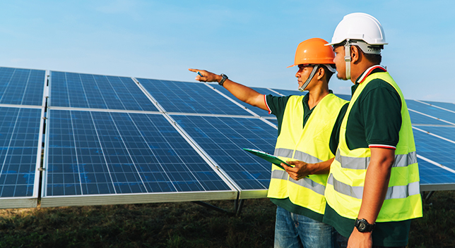 Dois trabalhadores de campo em coletes e capacetes em pé na frente de um grande painel solar e um dos trabalhadores está apontando