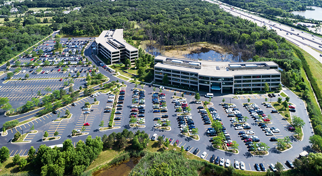 An aerial view of a corporate building and a parking lot full of cars built near a pond and wooded area with a freeway behind it