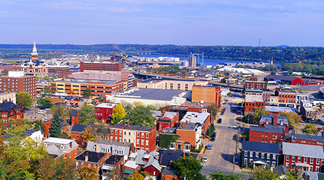 A landscape view of Dubuque, Iowa with bright colored homes, green trees, and factories on a waterfront
