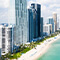 The beach coastline of Miami Beach, Florida with tall skyscrapers and people out on the white sand beaches next to the clear blue water