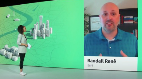 A screencap from the featured video showing a casually-dressed person looking at a large wall display with a computer model of a city in white on a green background and a photo of Randall René