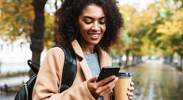 Photo d’une personne portant un manteau rose pâle et tenant une tasse de café et souriant en se penchant vers un téléphone portable qu’elle tient dans la main, se tenant dans une rue bordée d’arbres ensoleillée