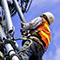 A photo of a utility worker in an orange vest and hard hat climbing a cellular base station to service the equipment against a cloud-streaked blue sky