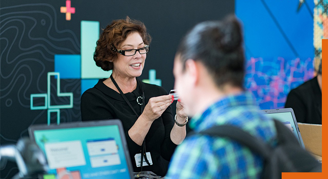 A photo of a conference staffer helping a registrant, while a person in a backpack stands in the foreground using a laptop