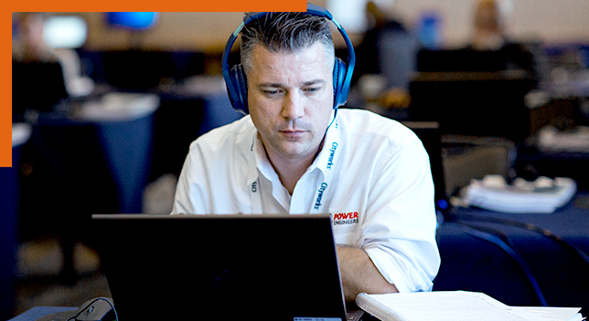 A photo of a person in a white collared shirt and a headset sitting in a large training room facing an open laptop with an expression of concentration