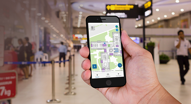 A hand holding a smartphone displaying an indoor map with the inside of a brightly lit, modern building in the background