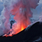 An erupting volcano showing bright orange lava spewing from two vents and red-glowing smoke plumes rising above a snowy mountain range