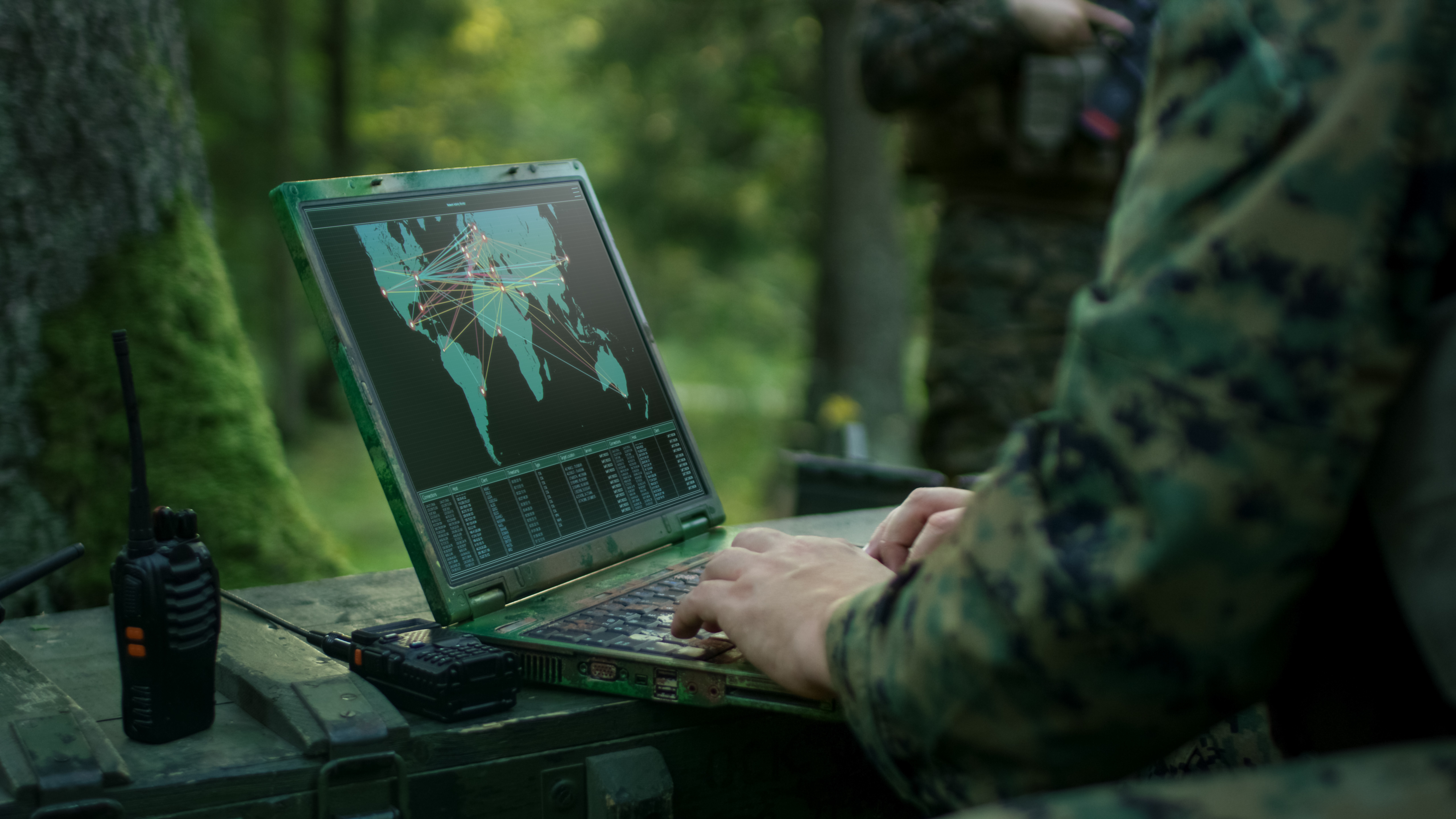 A person in green military fatigues accessing a map dashboard on an open laptop in a shady green forest