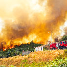 A wildfire raging on a wooded hillside with a red fire engine parked beside a small white house in the foreground
