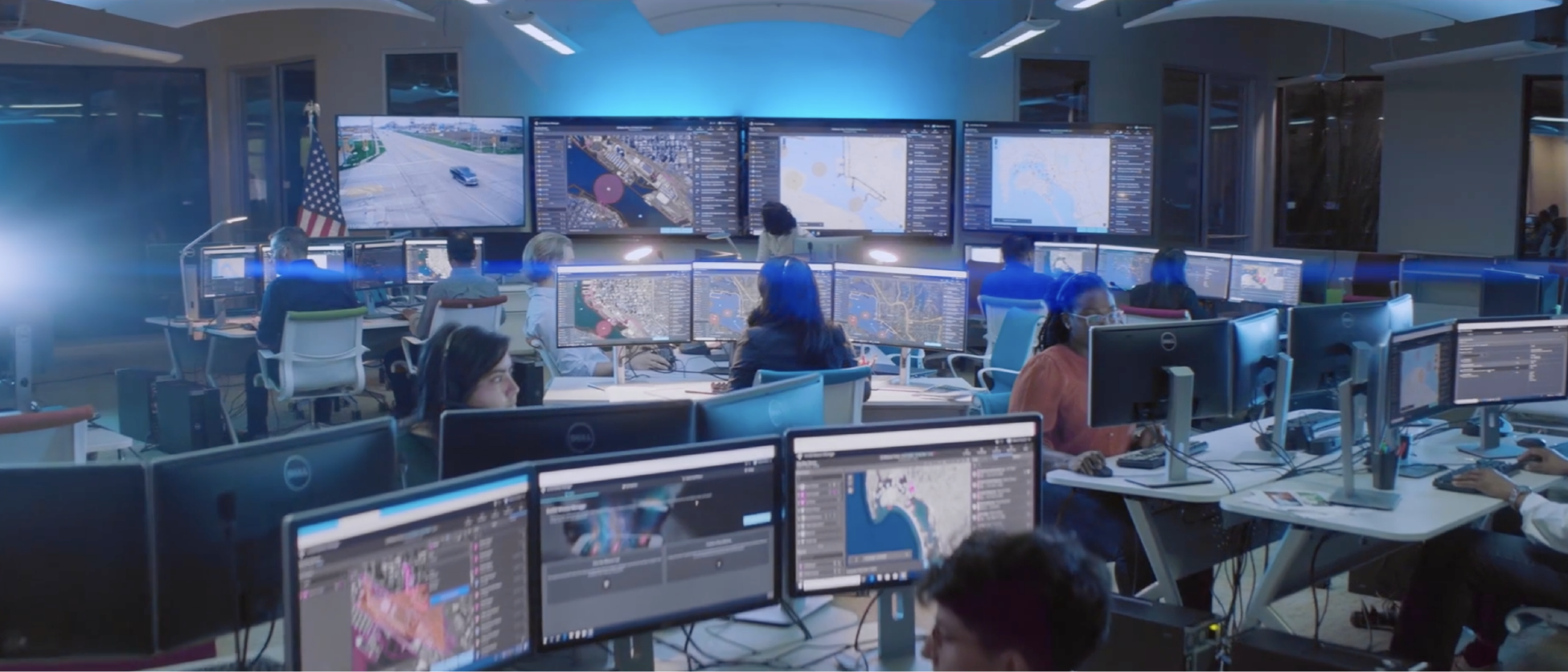 A blue-lit mission control room lined with large monitors displaying map dashboard data; and many people wearing headsets and sitting at computer stations