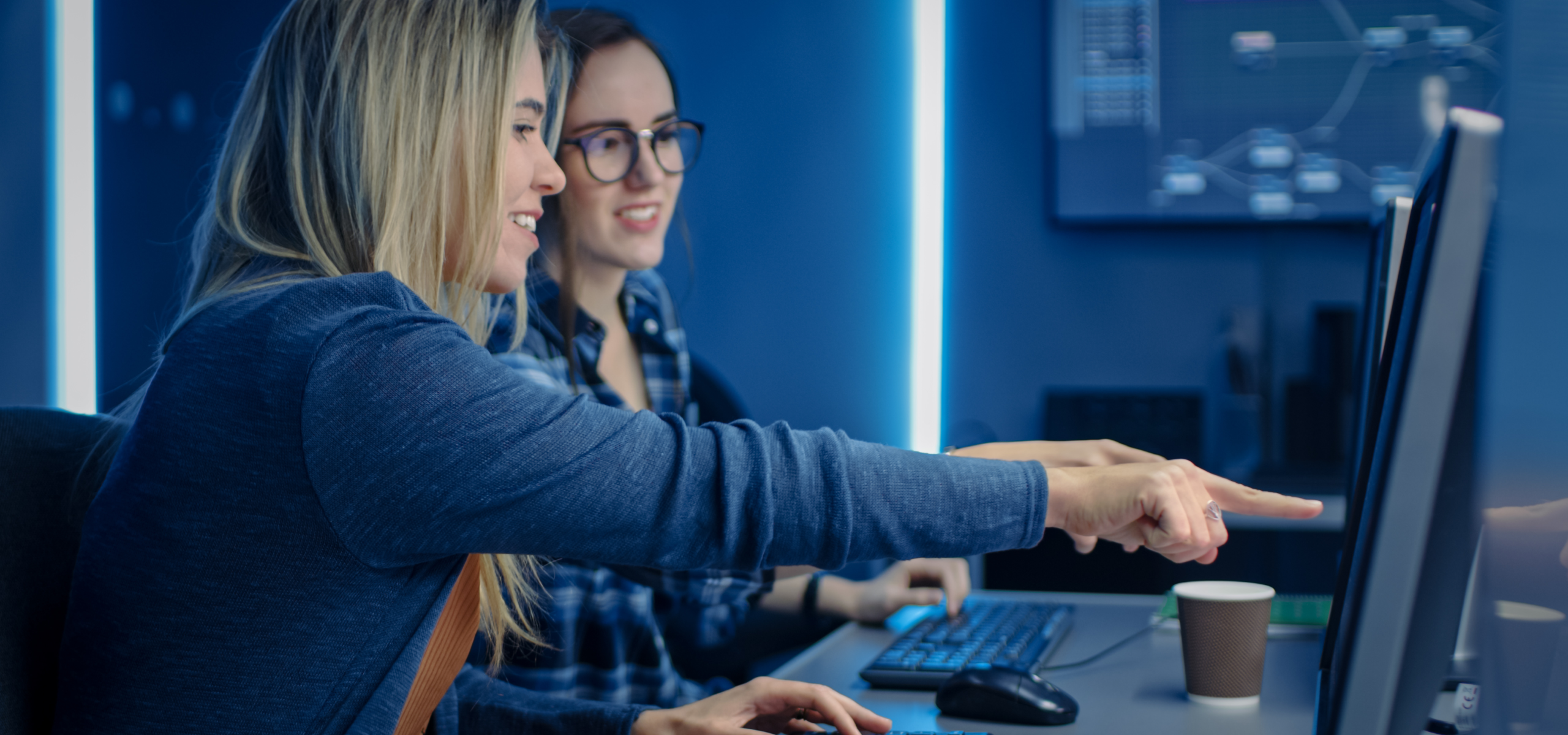 Two colleagues smiling as they discuss an unseen monitor display in a modern office setting dimly lit in shades of blue