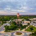 An aerial image of a water tower printed with the words “Round Rock” in a tree-filled city square under a wide overcast sunrise sky of blue and pale orange
