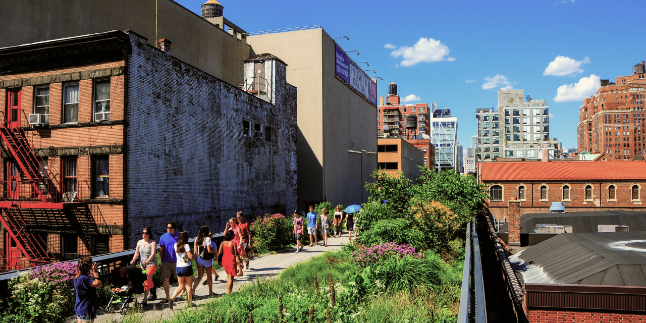 Several people in warm-weather attire walk along part of the High Line in New York City with lush greenery and blooming flowers.