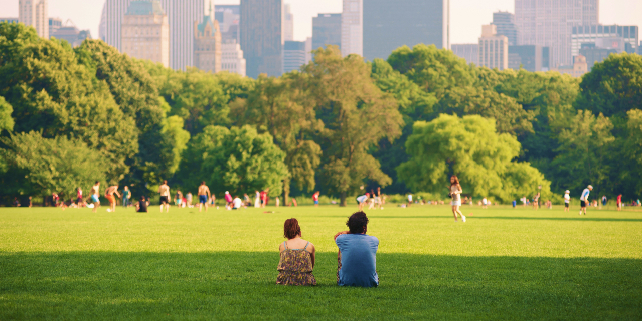 A young woman and man sit on a green grass field in Central Park facing away from the camera, watching others play soccer.
