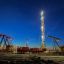 An oil drilling rig light brightly against approaching nighttime with a pump jack in motion in the foreground under a clear dark blue sky.