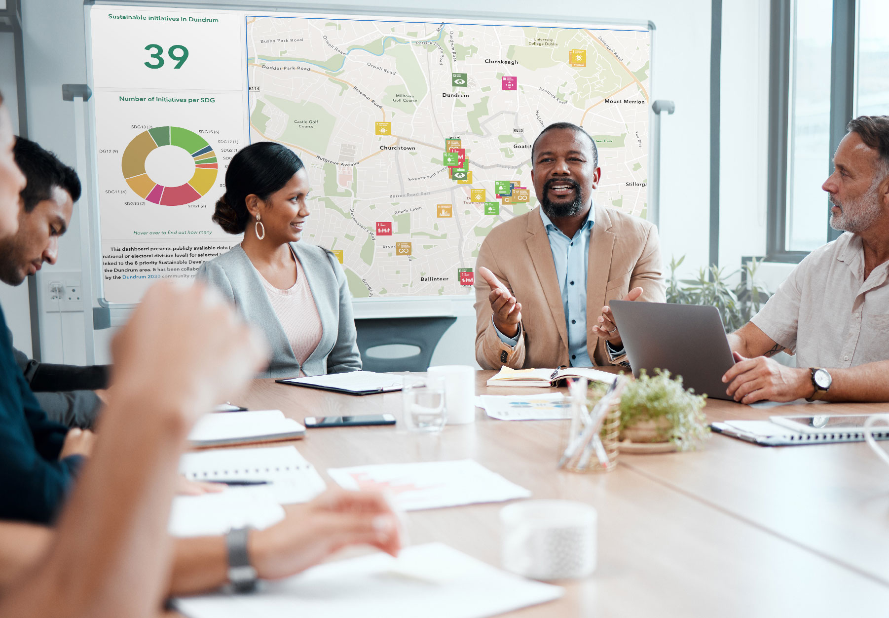Multiple people sitting around a conference table and talking with a GIS dashboard visible in the background
