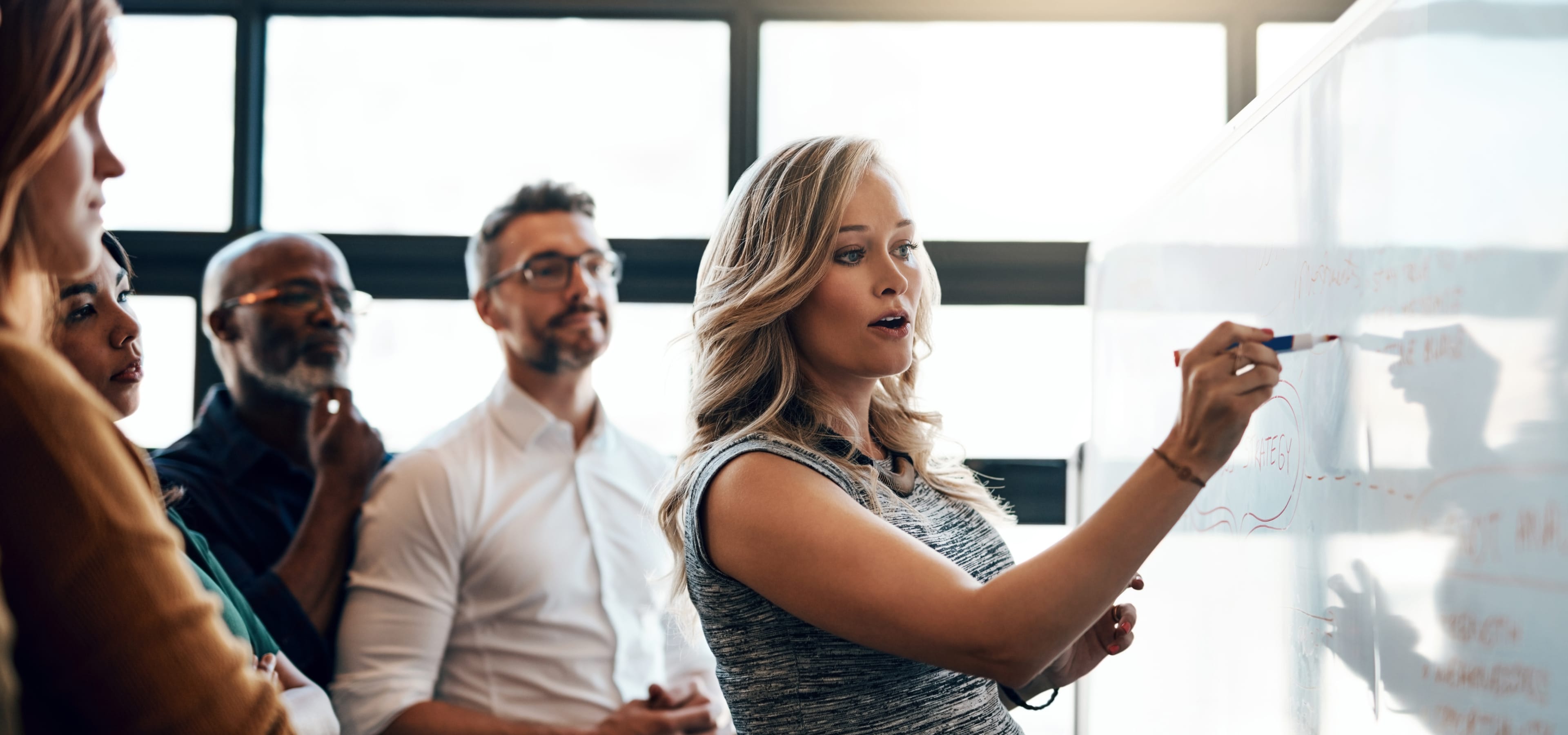 One woman writes on a whiteboard as four other professionals watch her attentively