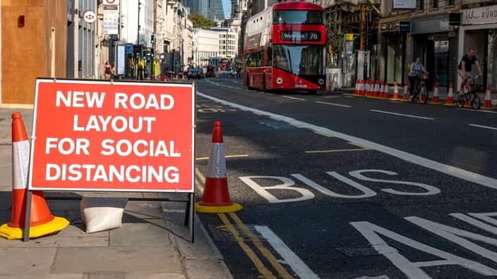 A street in London, United Kingdom features a bus lane and a sign on the sidewalk that reads “New Road Layout for Social Distancing”