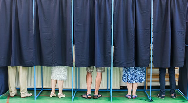 Five people’s feet are visible beneath closed curtains in adjacent voting booths