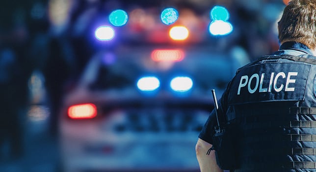 The back of a police officer wearing a vest that says “Police” is in focus in front of a police car in the background
