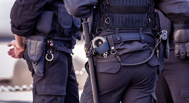 Three uniformed police officers stand together, focusing on one’s belt with a radio, handcuffs, and baton