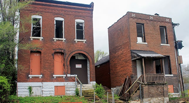 Two brick buildings in disrepair with boarded or broken windows and overgrown landscaping